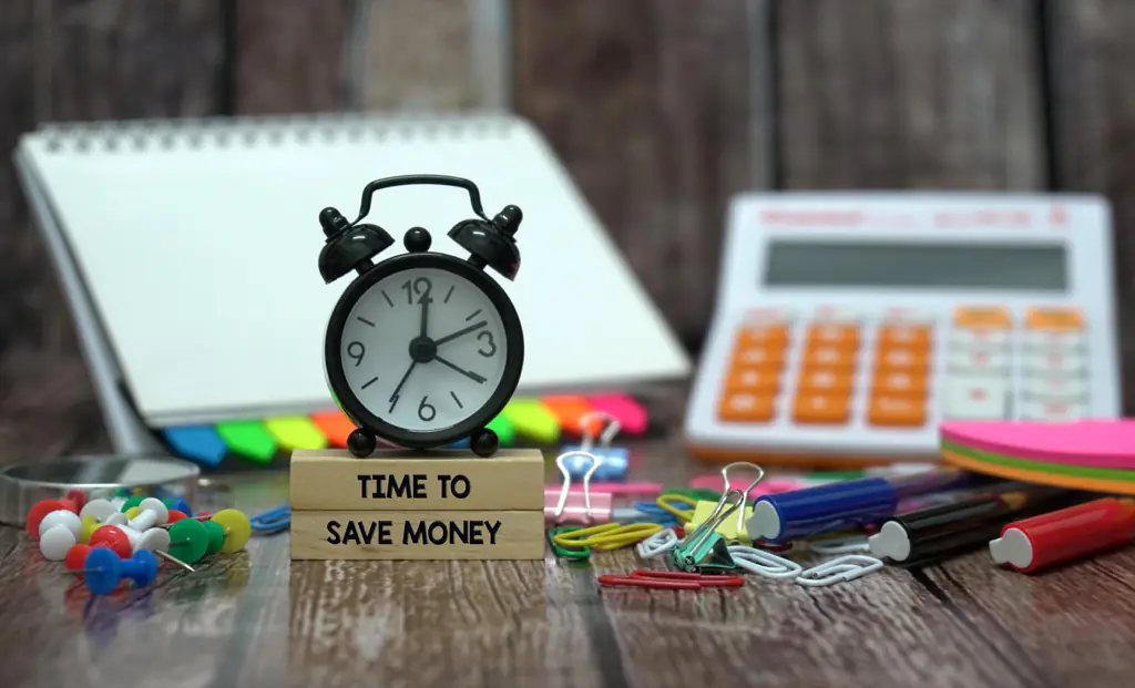 Desk with various office supplies and a clock on top of wooden blocks that have "Time to Save Money" imprinted.