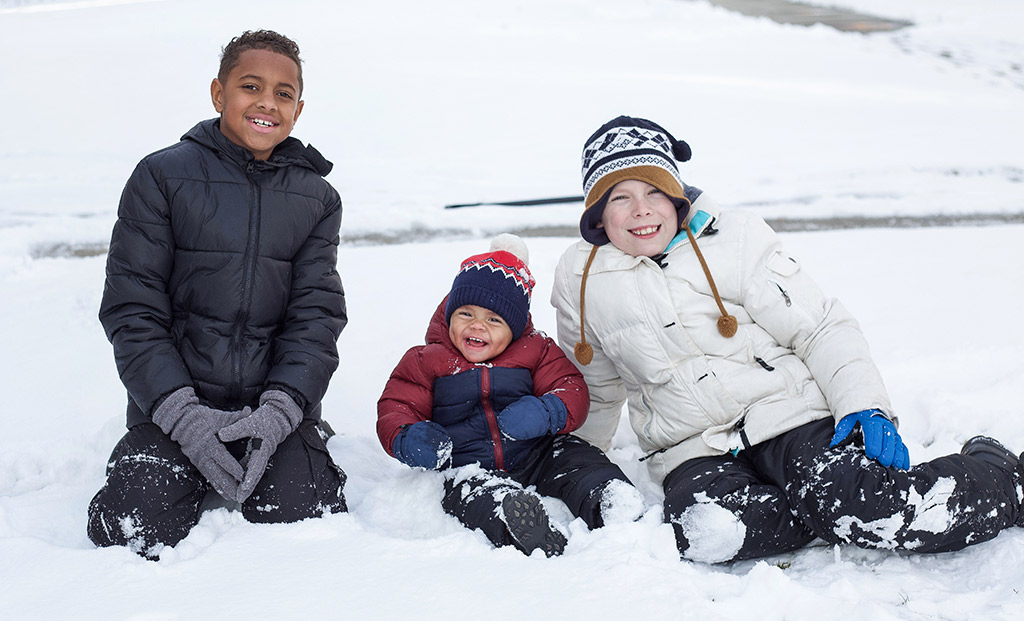 Three kids in winter clothes sitting in the snow.