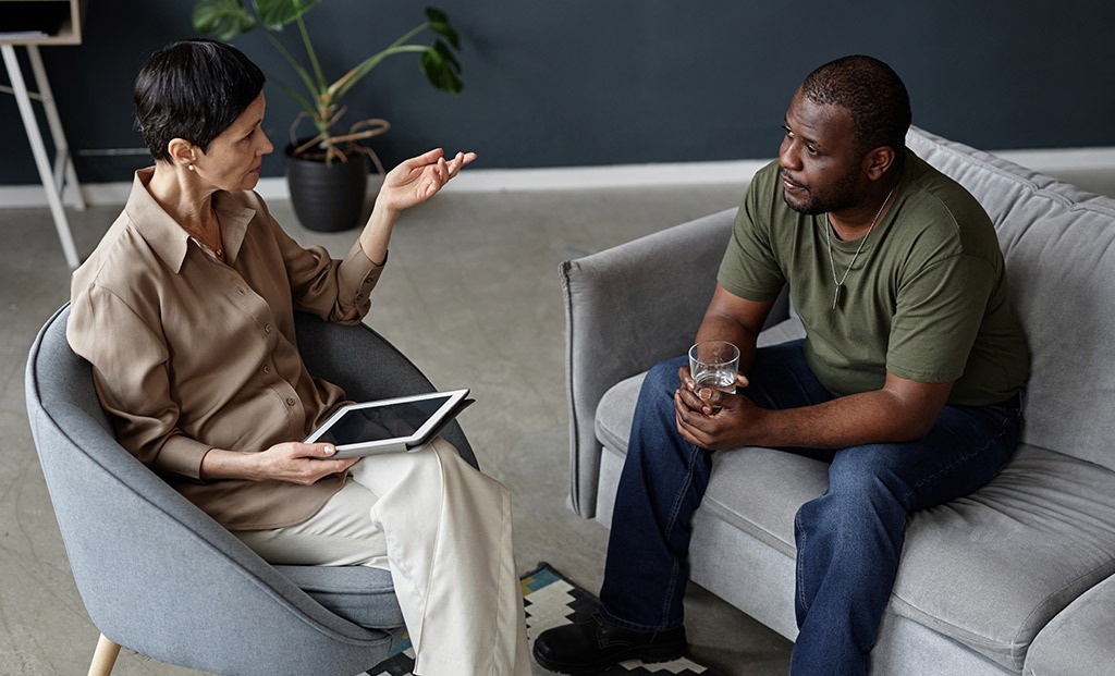 Veteran sitting on a couch with a glass of water having a conversation with a psychiatrist seated in a chair to the left